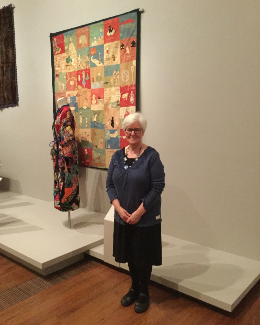 A woman stands in front of a colourful quilt hung on the wall of an art gallery